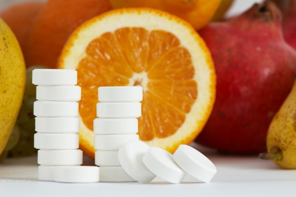 C-vitaminok egymásra halmozva, mögöttük félbevágott narancs és különféle gyümölcsök: körte, gránátalma.