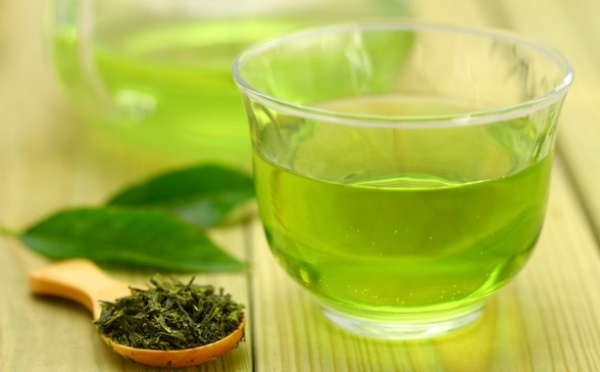 Üveg pohárban zöld tea, mellette szárított és friss tealevelek.
