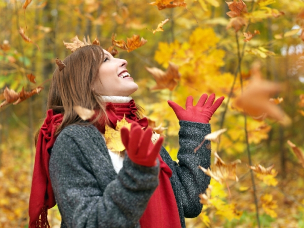 Piros kesztyűben és ugyanilyen színű sálban, meleg pulóverben egy fiatal lány mosolyogva nézi a lehulló faleveleket az őszi erdőben.