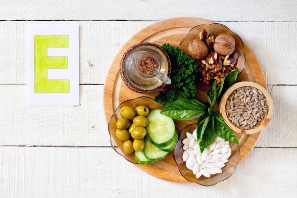 E-vitamint tartalmazó élelmiszerek összegyűjtve egy fából készült tálcán, mellette egy nagy sárga E betű.