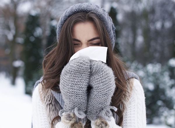 Orrát fújja egy szürke kesztyűt, sálat és sapkát viselő fiatal lány kinn, a szabadban, egy havas vidéken.