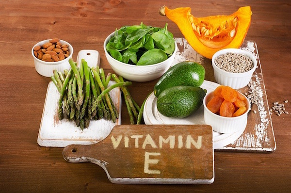 E-vitamin-tartalmú élelmiszerek egy tálcán: spárga, avokádó, aszalt sárgabarack, spenót, mandula, sütőtök.