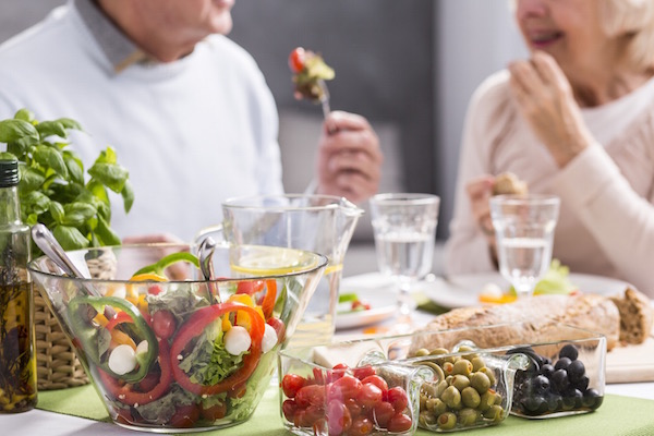 Idős házaspár asztalán csupa egészséges étel: friss saláták, zöldségek, olívabogyó, teljes kiőrlésű kenyér, olívaolaj és friss bazsalikom.