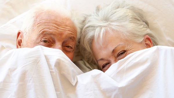 Idős házaspár az ágyban mosolyogva, akik a takarót magukra húzzák.
