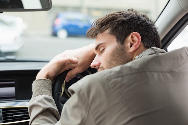 Fiatal férfi fáradtan ráborul az autó kormányára, még a szemét is behunyja.