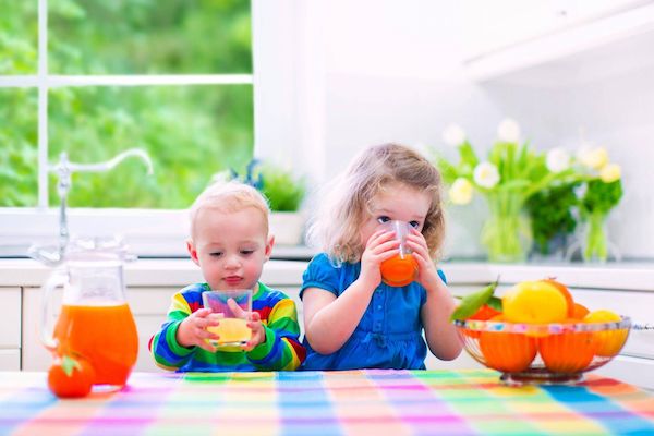 Két kisgyerek frissen facsart narancslevet iszik a konyhában, előttük egy tál magas C-vitamin-tartalmú gyümölcs.