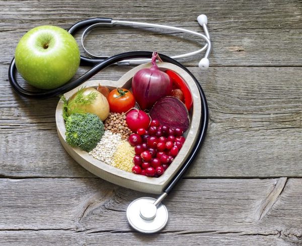 Szív alakú tálban egészséges zöldségek, gyümölcsök és gabonafélék, mellette egy fonendoszkóp és egy zöldalma.