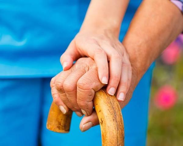Parkinson-kóros idős ember keze a botján, rajta a segítőjéé is.