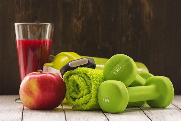 Piros gyümölcsökből egy pohár smoothie, alma, zöld kézisúlyzók és almazöld törülköző egymás mellett.