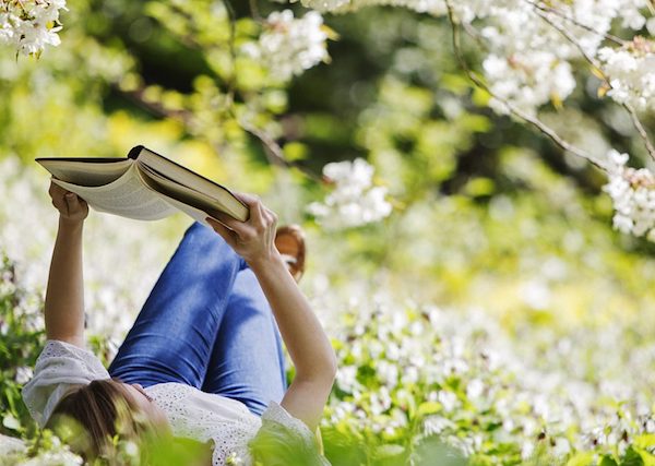 Virágzó fák alatt a fűben fekve olvas egy könyvet egy hölgy.
