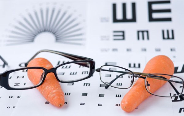 Vicces kép, melyen a látás fontosságát szimbolizálják két sárgarépára helyezett szemüveggel, a háttérben látásvizsgálatnál hazsnálatos ábrákkal.