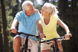 Idősebb házaspár szorosan egymás mellett biciklizik, mosolyognak egymásra.