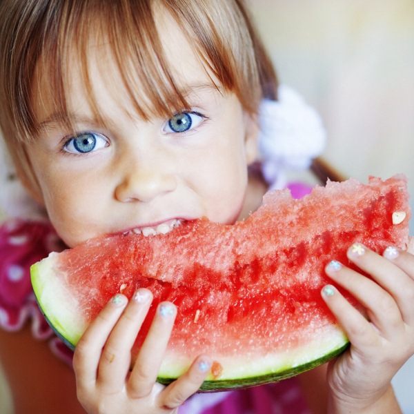 Kék szemű kislány egy nagy szelet görögdinnyét majszol.