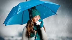 Sapkát és kesztyűt viselő lány orrát törli szakadó esőben egy kék esernyő alatt.