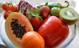 Sok C-vitamint tartalmazó zöldségek és gyümölcsök üvegtálon: kivi, piros paprika, eper, paradicsom, kelbimbó, narancs, lila káposzta és egy egzotikus gyümölcs félbevágva.