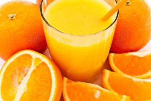 Félbevágott és egész narancsok, narancsszeletkék a képen, középen a gyümölcsből készült frissen préselt narancslé üvegpohárban, benne narancssárga szívószál.