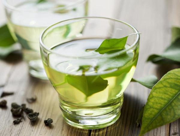 Üvegpohárban zöld tea, benne egy friss tealevéllel, mellette szárított tea.