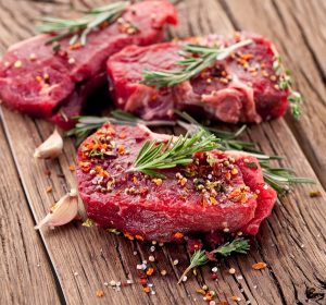 Szeletelt húsok meghintve fűszerekkel egy rusztikus faasztalon, mellettük fokhagyma, a húsokon rozmaringágacskák.