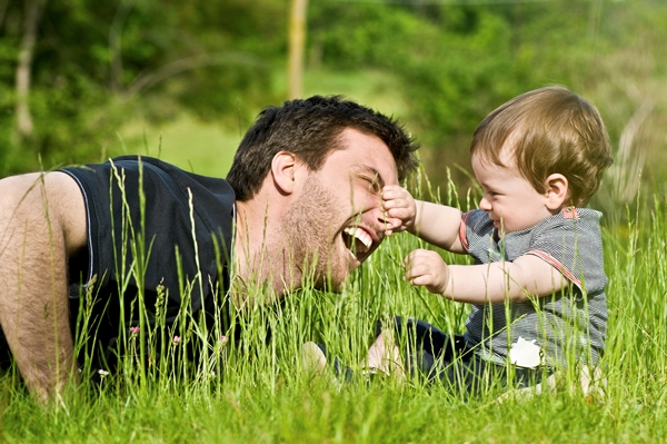 Fiatal férfi játszik kisfiával a réten a fű között, a baba épp apukája orrát markolássza, aki nevet közben.