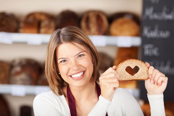 Pékségben egy mosolygós eladó egy teljes kiőrlésű kenyér szeletkéjét tartja a kezében, melyből kivágtak egy szív alakot.