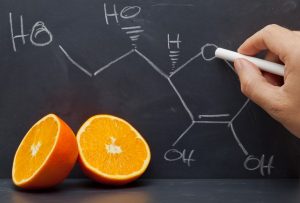 A C-vitamin kémiai képletének felírása fehér krétával egy fekete táblára, az asztalon egy félbevágott narancs mint C-vitamin-forrás.