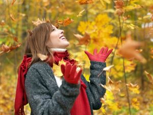 Piros kesztyűben és ugyanilyen színű sálban, meleg pulóverben egy fiatal lány mosolyogva nézi a lehulló faleveleket az őszi erdőben.