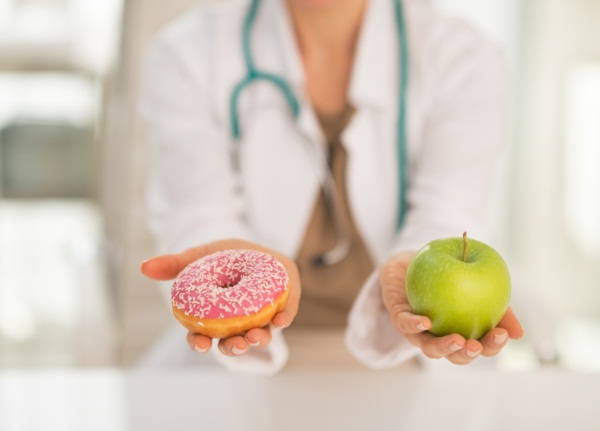 Egyik kezében cukros fánkot, másikban egy zöld almát tart egy doktornő, kinek fonendoszkóp lóg a nyakában.