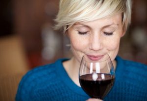 Szőke hajú nő kék pulóverben egy pohár vörösbort készül inni.