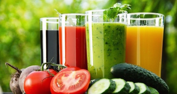 Frissen facsart színes zöldség- és gyümölcslevek, mellettük az alapanyagok: cékla, paradicsom és uborka.