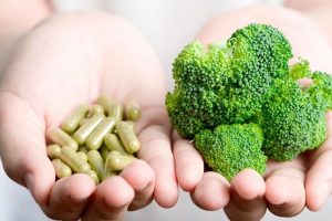 Két tenyér egymás mellé téve, egyikben a friss vitamint szimbolizáló brokkoli, másikban zöld étrend-kiegészítő kapszulák.