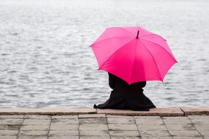 Egyedül ül az esőben a folyó rakpartján egy fekete kabátos nő, kezében pink esernyő.