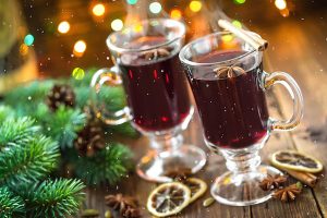 Karácsonyi hangulatú kép fenyőággal, tobozokkal, fényekkel és két pohár gőzölgő forralt borral.