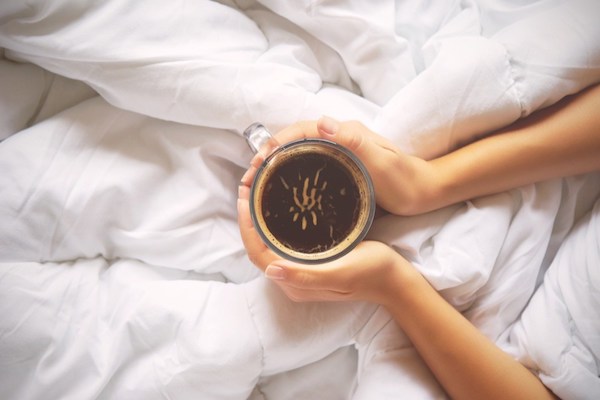 Reggeli kávéját fogja két kezébe egy fiatal lány az ágyban.
