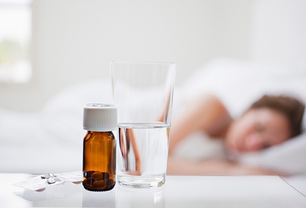 Alvó nő ágya mellett egy pohár víz és különféle vitaminok, kapszulák.