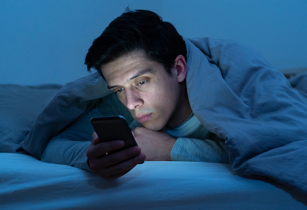 Mobiltelefonján néz valamit egy férfi az ágyábáan.