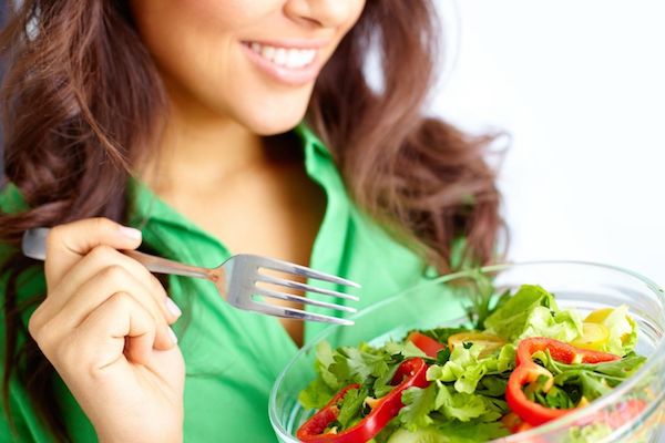 Zöld blúzos hölgy friss salátával teli tálból eszik, kezében villával.