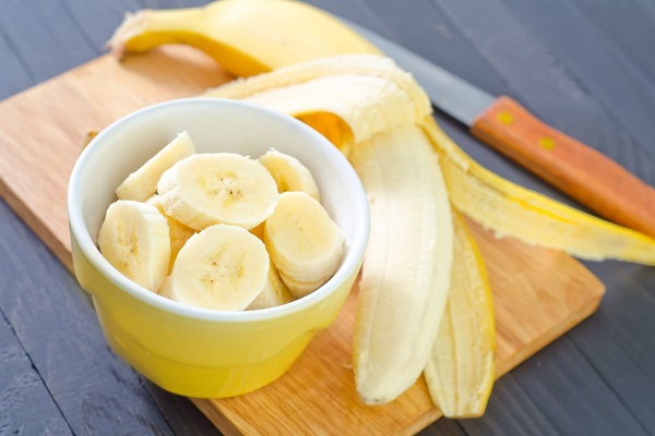 Vágódeszkára helyezett tálban banánszeletek, mellette egy banán és kés.
