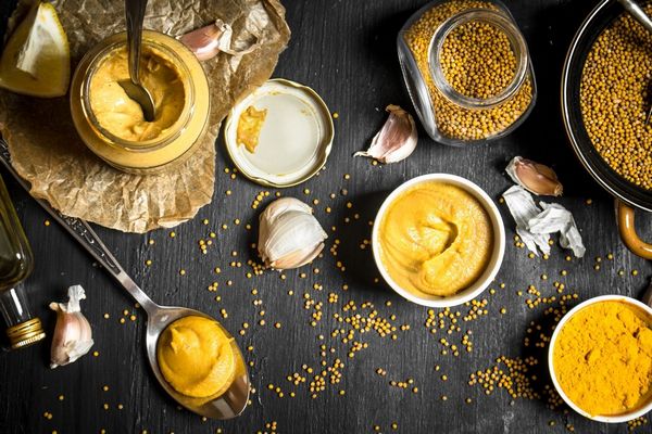 Egy szürke asztalon mustármagok kis üvegben és egy tálban, mellettük tálakban és kanálban mustár és fokhagymagerezdek, illetve olaj és citromszelet.