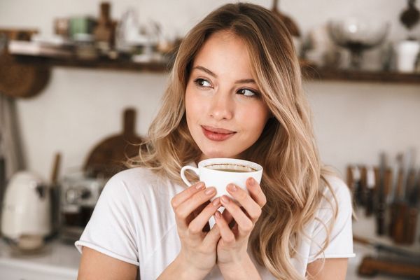 Fiatal mosolygós nő egy kávézóban két kezével kávéscsészét tart, miközben oldalra néz.