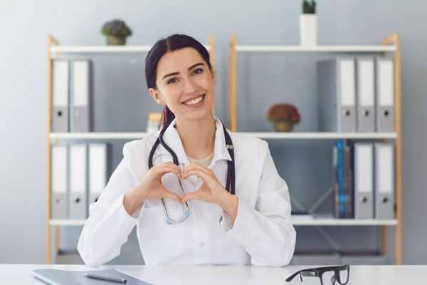 Egy orvosi rendelőben egy mosolygós doktornő fonendoszkóppal a nyakában két kezével szívet mutat.