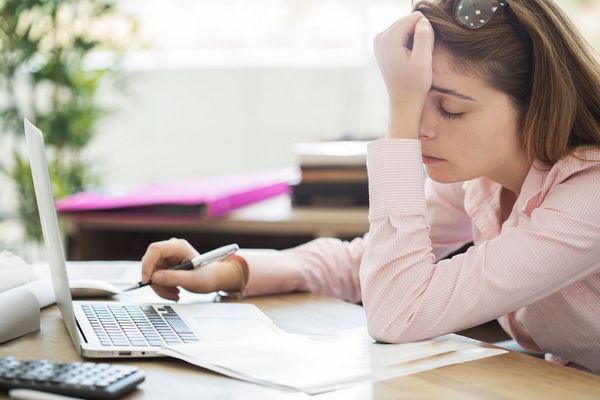 Egy fiatal nő az irodában laptoppal dolgozik, kezével a fáradt fejét tartja, miközben megpihen.