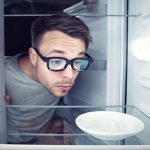 Egy szemüveges fiatal férfi benéz egy üres hűtőbe, ahol egy üres tányért néz.