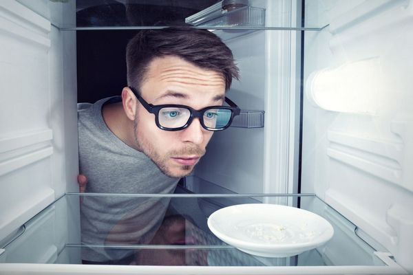 Egy szemüveges fiatal férfi benéz egy üres hűtőbe, ahol egy üres tányért néz.