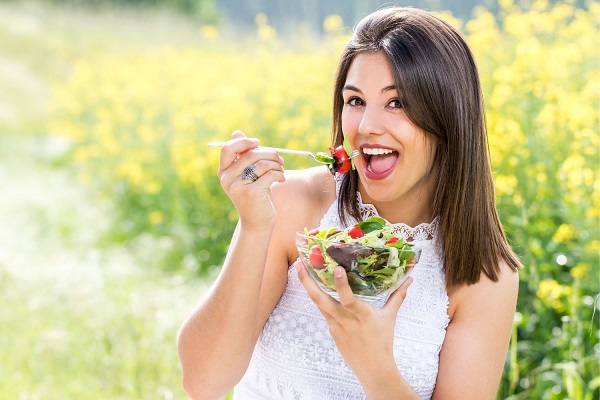 Egy fiatal nő virágos réten ül, kezében salátástál, salátát eszik.