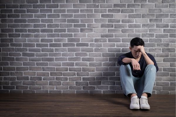 Egy fiatal férfi depressziósan ül egy szürke téglás fal előtt a földön.