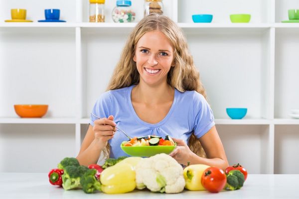 Egy fiatal lány a konyhában salátát eszik, körülötte sok zöldség, paradicsom, paprika, karfiol, brokkoli.
