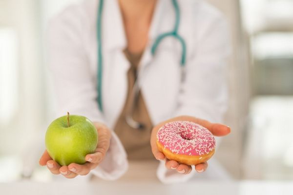Egy fehér köpenyes orvos egyik kezében egy alma, a másik kezében donat fánk.