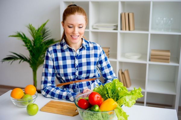 Egy fiatal nő kék-fehér kockás ruhában a konyhában ül, előtt salátástál és gyümölcsök, narancsok, almák, saláta, paradicsom, paprika, uborka.