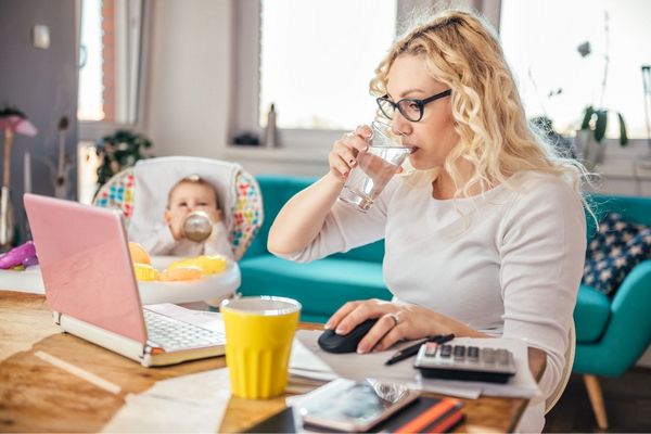 Egy fiatal anyuka home office-ban dolgozik, vizet iszik, mellette gyerekülésben a kisgyermeke cumisüvegből iszik.