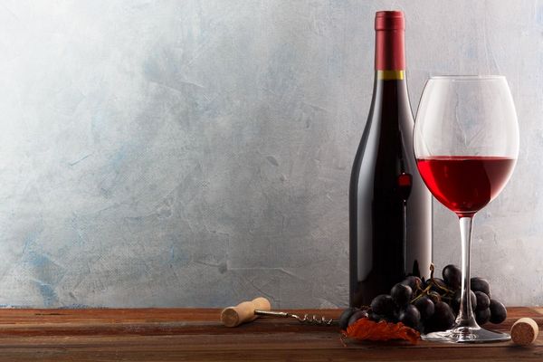 Egy asztalon vörösbor egy üvegben és egy borospohárban, mellette fekete szőlő és dugóhúzó.